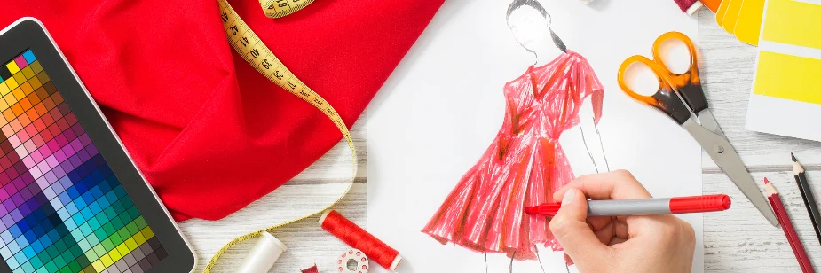 A imagem mostra uma mão desenhando um croqui de moda, destacando um vestido vermelho. Ao redor, há diversos itens relacionados à costura e design, como um tecido vermelho, uma fita métrica amarela, uma tesoura laranja, carretéis de linha, lápis e uma paleta de cores. Também é visível um tablet com uma tabela de cores, sugerindo o uso de tecnologia no processo criativo.